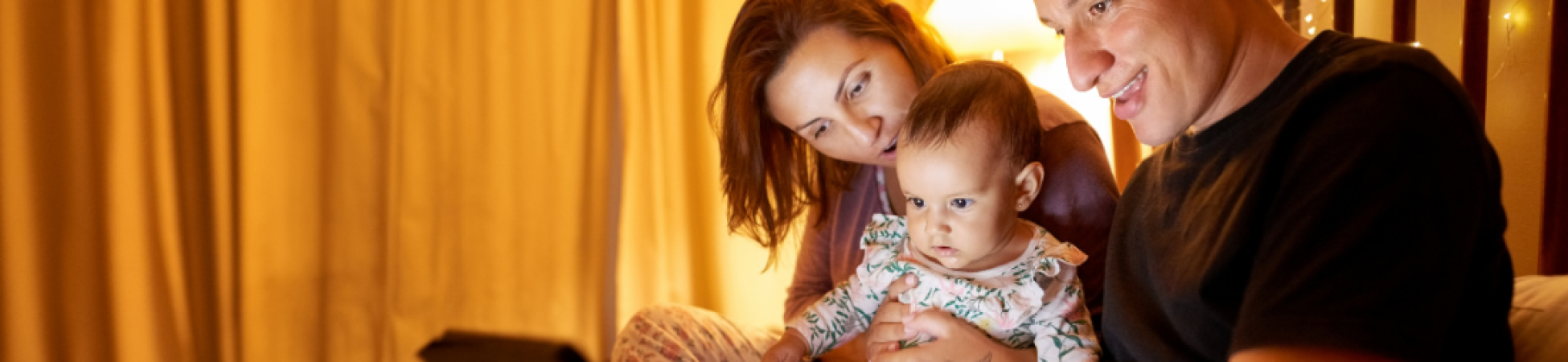 The photo highlights a warm family moment, with parents and child gathered around a device, likely enjoying a bedtime story. The soft lighting and cozy atmosphere create the perfect setting for bonding over this nightly tradition.